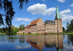 Das Schloss Raesfeld, welches aus einem Turm und mehreren Gebäuden, die im rechten Winkel zueinander stehen, besteht. Vor dem Schloss befindet sich ein See, in dem sich das Gebäude spiegelt.